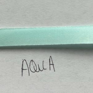 May include: A smooth, light aqua-colored satin ribbon strip positioned horizontally across the frame. Below the ribbon, the word "Aqua" is handwritten in black ink on a plain white surface, clearly labeling the specific color of the ribbon.