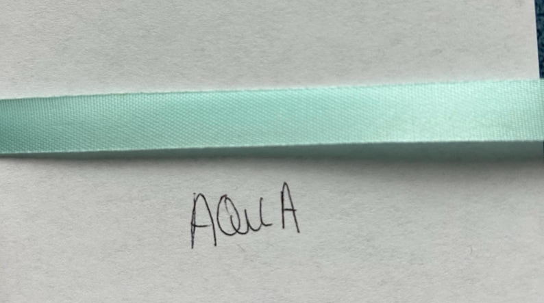 May include: A smooth, light aqua-colored satin ribbon strip positioned horizontally across the frame. Below the ribbon, the word "Aqua" is handwritten in black ink on a plain white surface, clearly labeling the specific color of the ribbon.