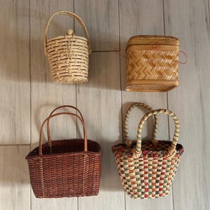 May include: Four woven baskets of varying shapes and sizes. The baskets are made of natural and dyed materials, with handles and lids. The baskets are in shades of brown, beige, and multi-colored patterns. The baskets are displayed against a light wood background.