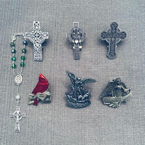 May include: An assortment of religious items including silver-toned crosses, a rosary with green beads, a red cardinal figurine, and two angel figurines. The crosses feature intricate designs and text. The cardinal figurine has a heart that says "Safe Travels".