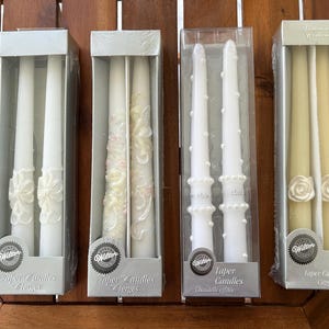 May include: Four boxes of taper candles in various designs. The candles are white and ivory, with decorative elements like bows and roses. The boxes are labeled "Taper Candles" and "Wilton".