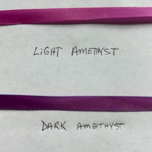 May include: Two horizontal strips of ribbon. The top ribbon is a light amethyst purple color with the handwritten text Light Amethyst below it. The bottom ribbon is a dark amethyst purple color with the handwritten text Dark Amethyst below it.