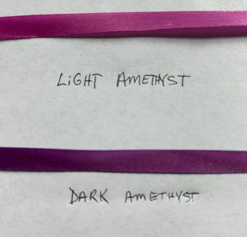 May include: Two horizontal strips of ribbon. The top ribbon is a light amethyst purple color with the handwritten text Light Amethyst below it. The bottom ribbon is a dark amethyst purple color with the handwritten text Dark Amethyst below it.