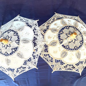 May include: Two white lace parasols with wooden handles. The parasols have intricate floral patterns and a delicate, openwork design. The background is a dark blue surface.