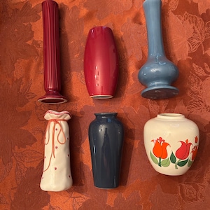May include: A collection of six ceramic vases in various shapes and colors. The vases include a tall, ribbed burgundy vase, a rounded red vase, a blue vase with a bulbous base, a white vase with a drawstring top, a dark blue vase, and a white vase with tulip designs.