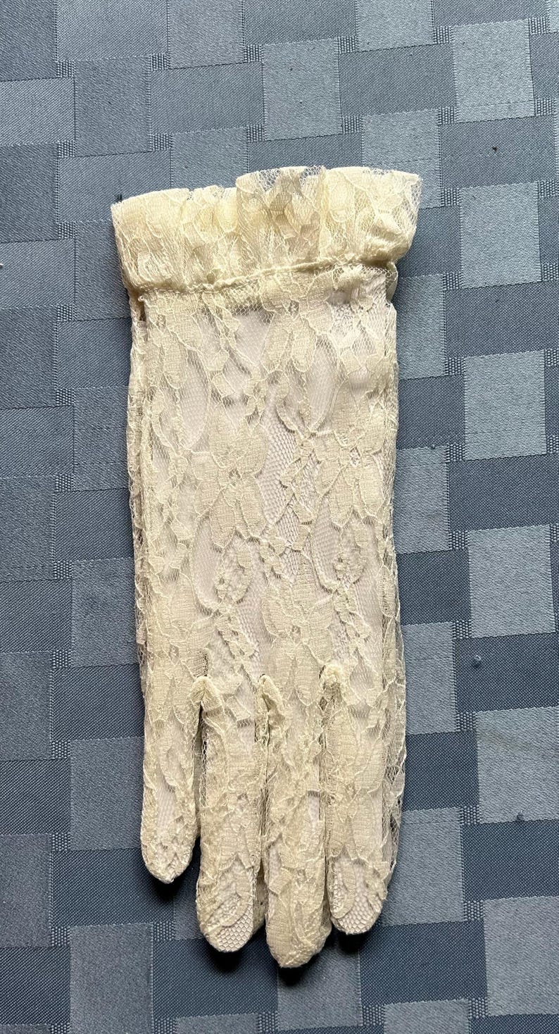 May include: A pair of ivory lace gloves with a delicate floral pattern. The gloves have a ruffled cuff and are made of a sheer fabric.