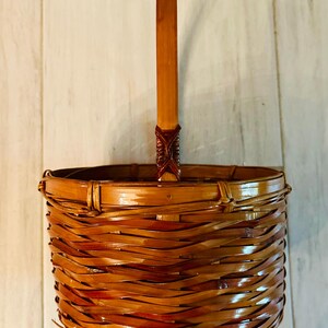 May include: A woven brown bamboo basket with a handle. The basket has a round shape with a handle that extends upwards. The basket is made of woven bamboo strips and has a glossy finish. The handle is attached to the basket with a decorative woven element.