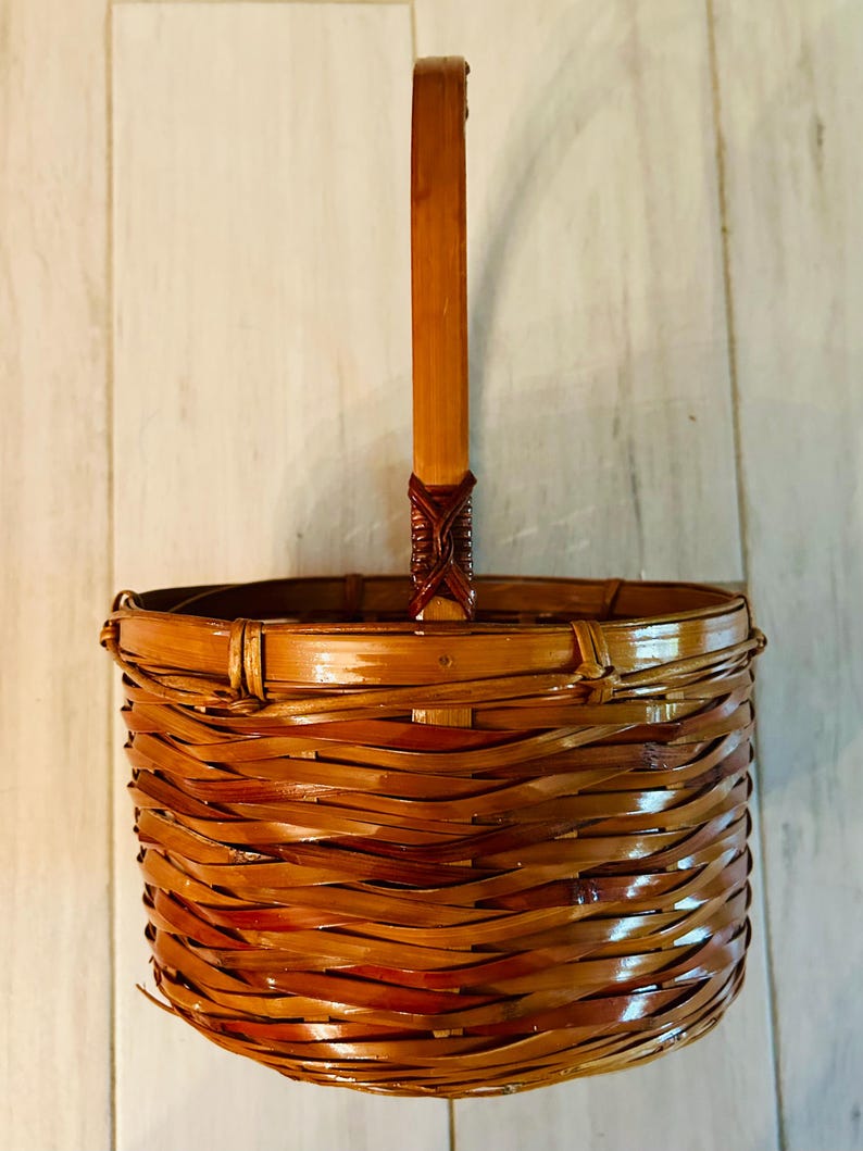 May include: A woven brown bamboo basket with a handle. The basket has a round shape with a handle that extends upwards. The basket is made of woven bamboo strips and has a glossy finish. The handle is attached to the basket with a decorative woven element.