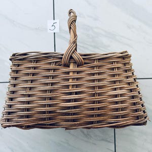 May include: A rectangular, woven wicker basket with a braided handle. The basket is light brown and has a square shape. The number "5" is written on a white square attached to the basket.