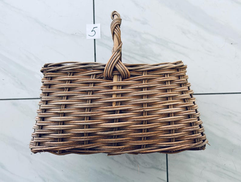 May include: A rectangular, woven wicker basket with a braided handle. The basket is light brown and has a square shape. The number "5" is written on a white square attached to the basket.