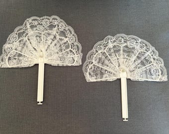May include: Two white hand fans featuring delicate lace fabric with a floral pattern and scalloped edges. Each fan is attached to a solid white plastic handle with a central pivot point, allowing the lace to spread into a semi-circular shape.