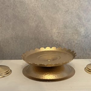 May include: Two gold-toned metal candle holders with slender stems and circular bases, positioned on either side of a central gold-toned metal pedestal tray featuring a scalloped edge and a textured, hammered finish on its surface.