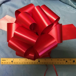 May include: A large, vibrant red satin bow with multiple loops and tails. A wooden ruler with inch measurements is placed next to the bow. The bow is likely for gift wrapping or decoration.