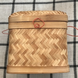May include: A woven bamboo box with a red string closure. The box is oval shaped and has a natural wood finish.