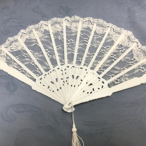 May include: A white lace hand fan with a white tassel. The fan is closed and has a delicate lace design.