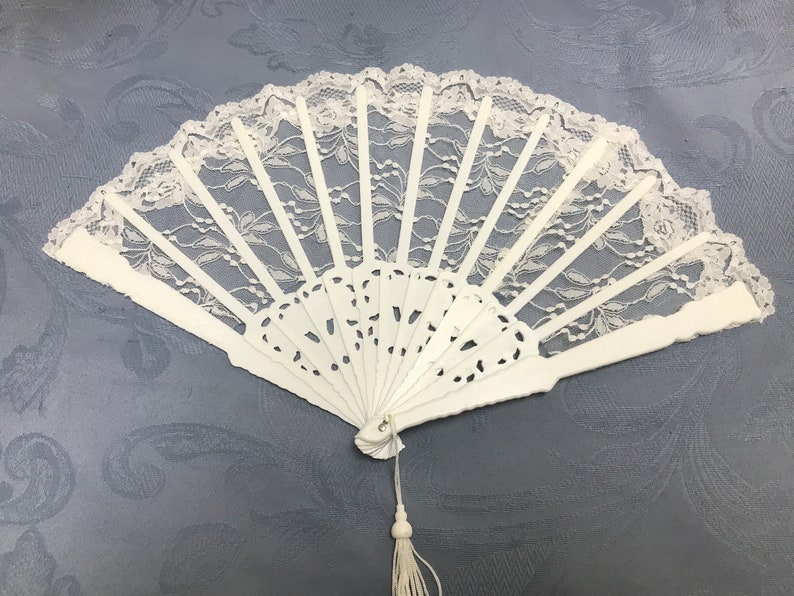 May include: A white lace hand fan with a white tassel. The fan is closed and has a delicate lace design.