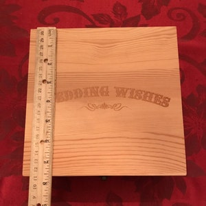 May include: A wooden box with a sliding lid. The box is light brown and has the words "Wedding Wishes" engraved on the front. A ruler is placed next to the box for size reference.