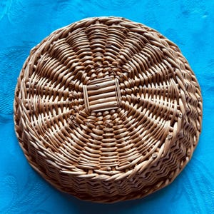 Puede incluir: Una cesta redonda de mimbre trenzado con un acabado natural de color marr&oacute;n claro. La base presenta un patr&oacute;n de tejido cuadrado distintivo en el centro, con filas circulares conc&eacute;ntricas de juncos trenzados que irradian hacia afuera para formar los lados poco profundos y texturizados.