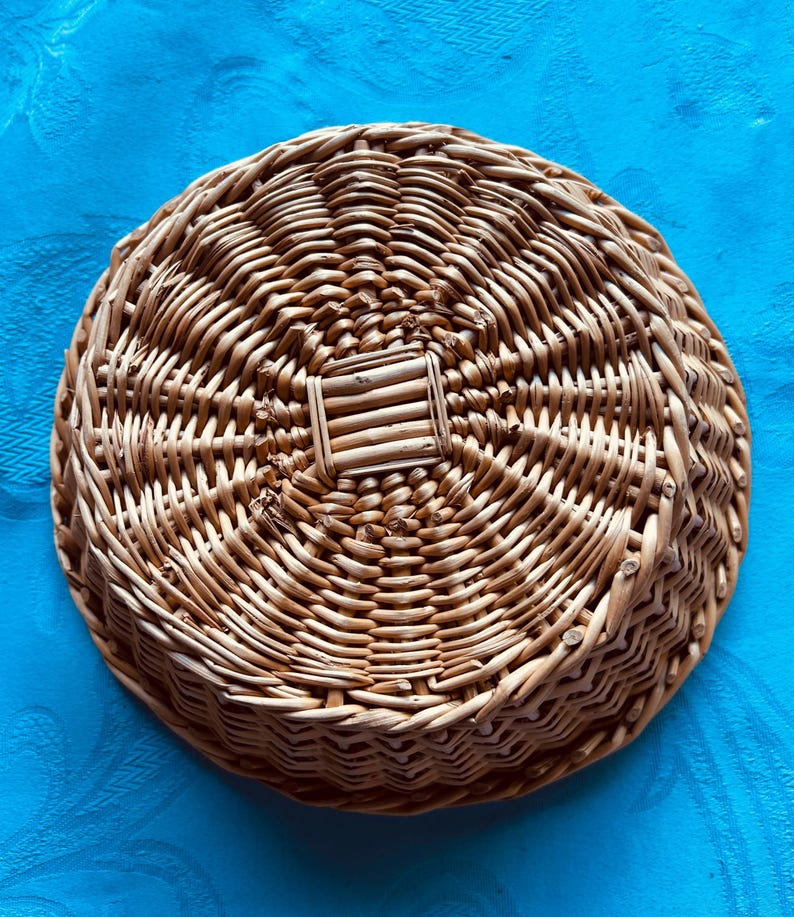 Puede incluir: Una cesta redonda de mimbre trenzado con un acabado natural de color marr&oacute;n claro. La base presenta un patr&oacute;n de tejido cuadrado distintivo en el centro, con filas circulares conc&eacute;ntricas de juncos trenzados que irradian hacia afuera para formar los lados poco profundos y texturizados.