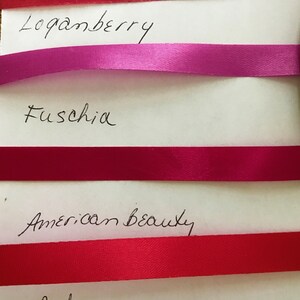 May include: Four horizontal satin ribbons in shades of Loganberry, Fuschia, American Beauty, and Red, each labeled with its corresponding color name in black handwritten text on a white background, showcasing a smooth, reflective texture.