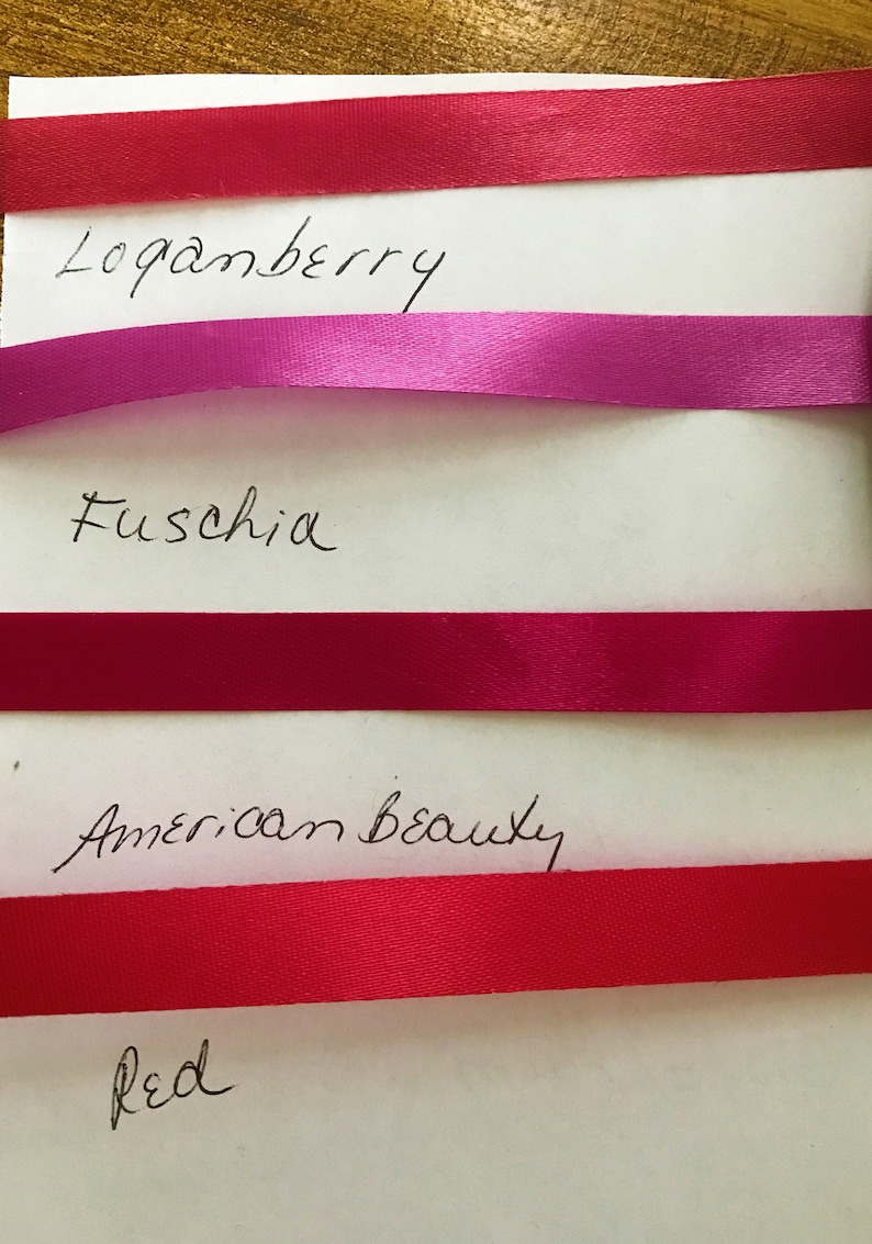 May include: Four horizontal satin ribbons in shades of Loganberry, Fuschia, American Beauty, and Red, each labeled with its corresponding color name in black handwritten text on a white background, showcasing a smooth, reflective texture.