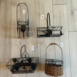 May include: Four black metal wall mounted baskets with woven straw bottoms. The baskets have a vine and leaf design. The baskets are numbered 1 through 4.