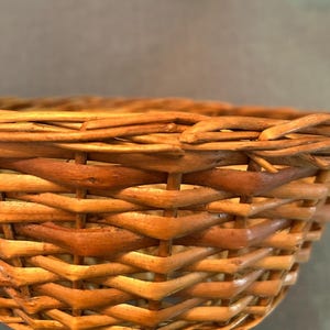 Puede incluir: Una cesta tejida hecha de juncos naturales de color marr&oacute;n claro, con un patr&oacute;n entrelazado detallado con hebras verticales y horizontales visibles. El borde est&aacute; construido con un junco m&aacute;s grueso y redondeado, mostrando la textura.