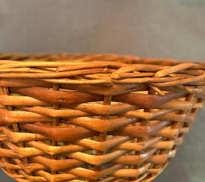 Puede incluir: Una cesta tejida hecha de juncos naturales de color marr&oacute;n claro, con un patr&oacute;n entrelazado detallado con hebras verticales y horizontales visibles. El borde est&aacute; construido con un junco m&aacute;s grueso y redondeado, mostrando la textura.