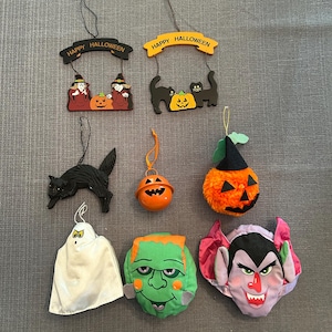 May include: A collection of Halloween-themed decorations, including hanging signs with "Happy Halloween" text, a black cat, a ghost, a pumpkin bell, a Frankenstein head, and a vampire head. The decorations are in various colors, including orange, black, white, and green.