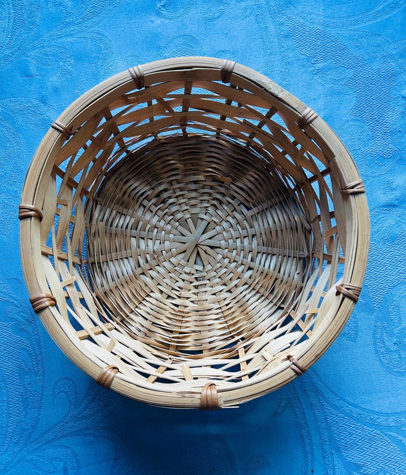 Puede incluir: Una cesta redonda de tejido abierto construida con tiras de bamb&uacute; natural de color marr&oacute;n claro. La cesta presenta una base circular con un patr&oacute;n de tejido radiante y lados verticales reforzados con una fina uni&oacute;n marr&oacute;n oscura.