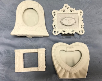 May include: Four white ceramic picture frames. One frame is shaped like a bell, another is a heart, and two are rectangular. The frame with the heart shape has a ruffle design. The rectangular frame with ornate details has the text "Especially For You" on a small tag.