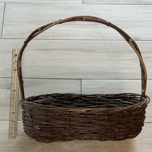 May include: A rustic, oval-shaped woven basket with a tall, arched handle. The basket is made of dark brown twigs and branches, with a ruler for scale. The handle is made of a thicker branch.