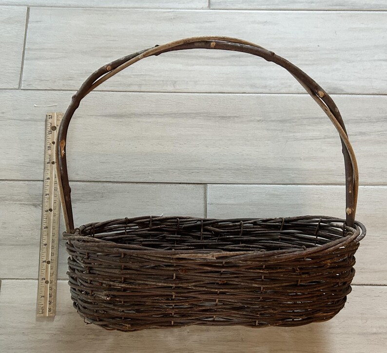 May include: A rustic, oval-shaped woven basket with a tall, arched handle. The basket is made of dark brown twigs and branches, with a ruler for scale. The handle is made of a thicker branch.