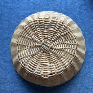 Puede incluir: Una cesta redonda de mimbre color beige claro con un patr&oacute;n radiante y texturizado. La cesta presenta un detalle de tejido cuadrado central que se transforma en una serie de crestas conc&eacute;ntricas repetitivas hacia el borde.