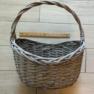 May include: A gray woven wicker basket with a handle. The basket is oval-shaped and has a sturdy handle. A wooden ruler is placed inside the basket. The basket is suitable for storage or display.