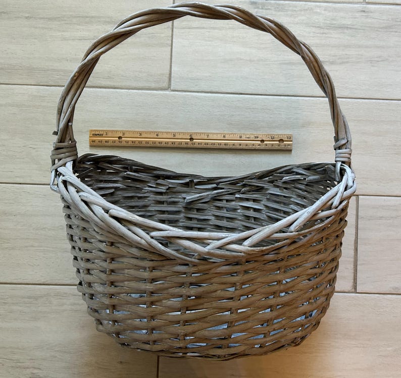 May include: A gray woven wicker basket with a handle. The basket is oval-shaped and has a sturdy handle. A wooden ruler is placed inside the basket. The basket is suitable for storage or display.