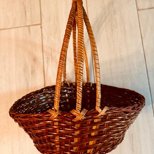 May include: A brown woven basket with a handle. The basket has a rounded shape with a wide opening and a woven design. The handle is made of three woven strands that meet at the top. The basket is made of a natural material.