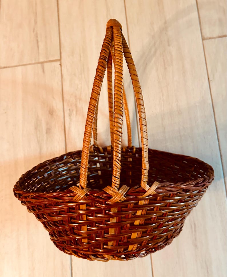 May include: A brown woven basket with a handle. The basket has a rounded shape with a wide opening and a woven design. The handle is made of three woven strands that meet at the top. The basket is made of a natural material.