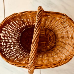 May include: Oval-shaped woven wicker basket with a tall, twisted handle. The basket is a warm brown color and has an open weave design. The handle is sturdy and provides a comfortable grip. This basket is suitable for storage or display.