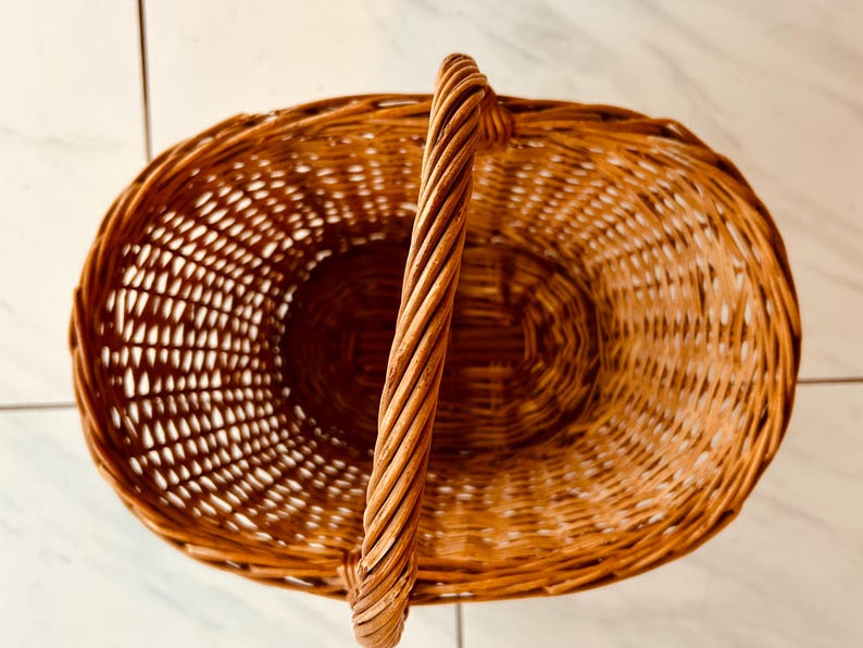 May include: Oval-shaped woven wicker basket with a tall, twisted handle. The basket is a warm brown color and has an open weave design. The handle is sturdy and provides a comfortable grip. This basket is suitable for storage or display.