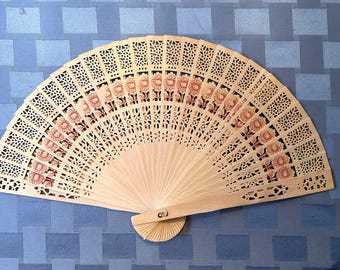 May include: A light beige wooden folding hand fan featuring intricate laser-cut geometric patterns and a repeating floral design in shades of pink and orange. The fan is fully extended, showcasing a delicate lattice structure and a central metal rivet.