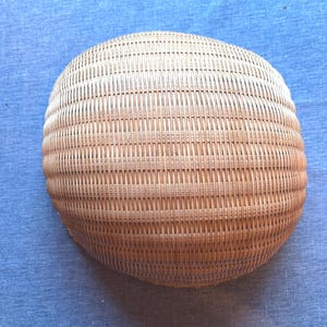 Puede incluir: Una cesta redonda y poco profunda de color madera natural, beige claro. La cesta est&aacute; construida con un patr&oacute;n de tejido apretado y uniforme de tiras finas verticales y horizontales, creando una superficie curva lisa con una textura consistente.