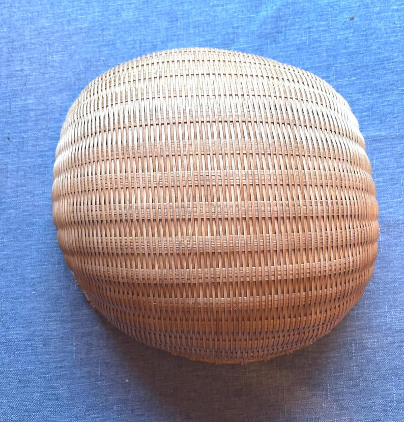 Puede incluir: Una cesta redonda y poco profunda de color madera natural, beige claro. La cesta est&aacute; construida con un patr&oacute;n de tejido apretado y uniforme de tiras finas verticales y horizontales, creando una superficie curva lisa con una textura consistente.