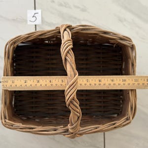 May include: Woven rectangular basket with a braided handle. A wooden ruler is placed across the top, measuring approximately 12 inches in length. The basket is made of light and dark brown wicker.