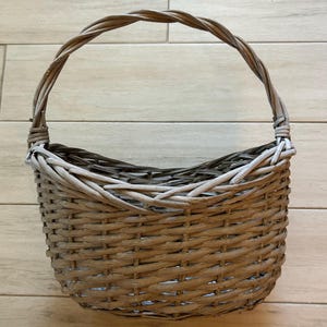 May include: A woven basket with a curved handle. The basket is made of light brown and gray wicker, with a rounded shape and a sturdy handle for carrying. The basket is empty and sits on a light brown wooden surface.