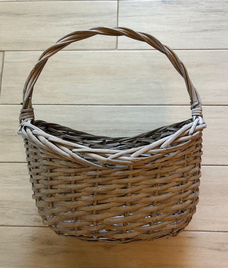 May include: A woven basket with a curved handle. The basket is made of light brown and gray wicker, with a rounded shape and a sturdy handle for carrying. The basket is empty and sits on a light brown wooden surface.