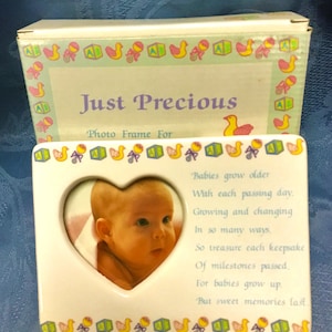 May include: A white ceramic photo frame with a heart-shaped opening, holding a baby's photo. The frame is decorated with baby-themed illustrations and the text "Just Precious." The frame includes a poem about cherishing baby milestones.