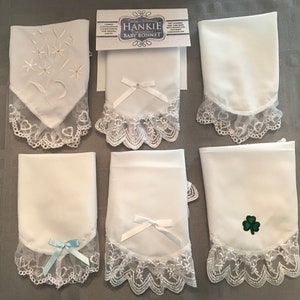 May include: Six white handkerchiefs with lace trim.  Each handkerchief has a different embroidered design, including a floral design, a shamrock, and a bow.  The handkerchiefs are packaged with a label that reads "HANKIE Embroidered Baby Bonnet".
