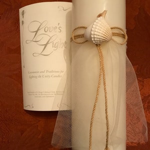 May include: A white pillar candle decorated with seashells, twine, and a sheer white fabric. The candle is accompanied by a card with the text "Love's Light" and information about unity candle ceremonies.