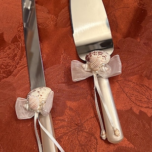May include: A set of two white handled cake knives with silver blades. The handles are decorated with a white bow and a seashell.